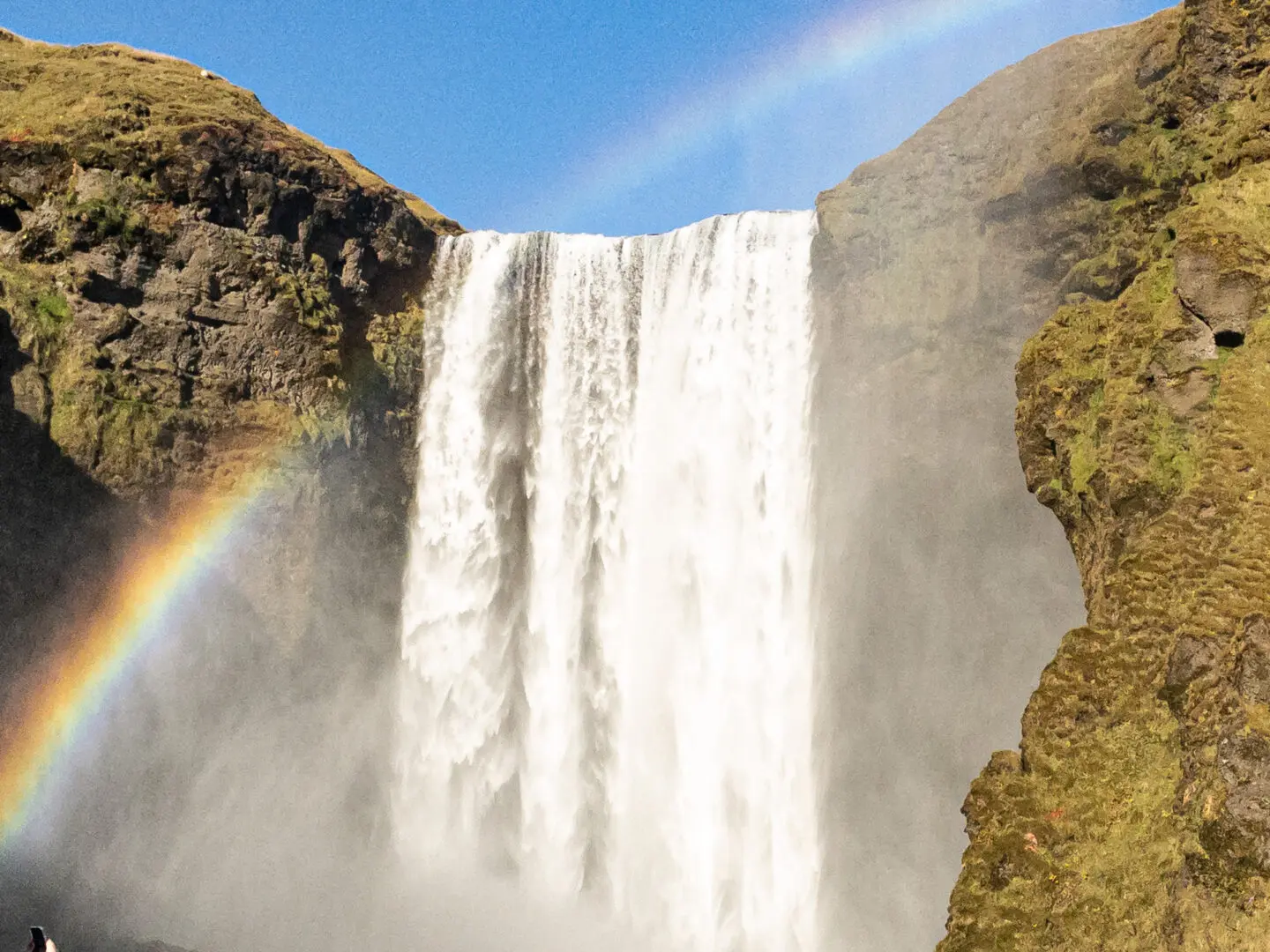 A rainbow is seen in the sky above a waterfall.